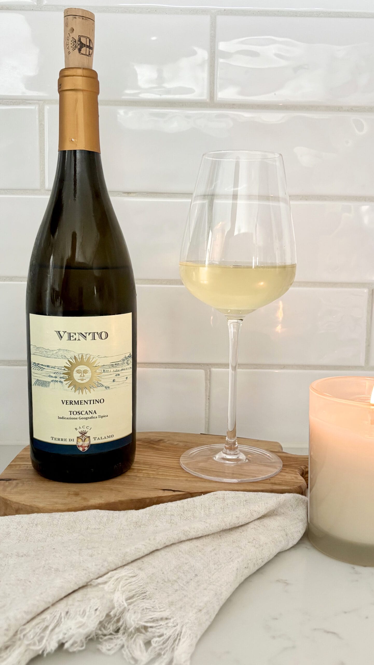 Bottle of Vento Vermentino Toscana beside a glass of white wine on a wooden board with linen cloth and candle against a white tile backsplash.