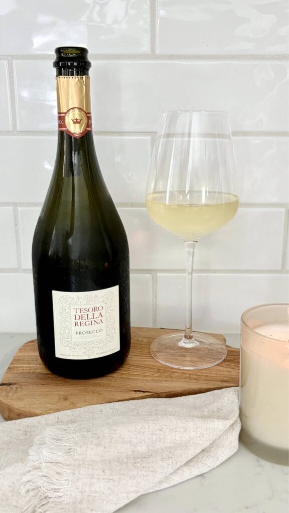 A chilled bottle of Prosecco beside a filled wine glass on an olive wood board. A lit candle and linen napkin create a simple Italian-inspired table setting against a white tile backsplash.