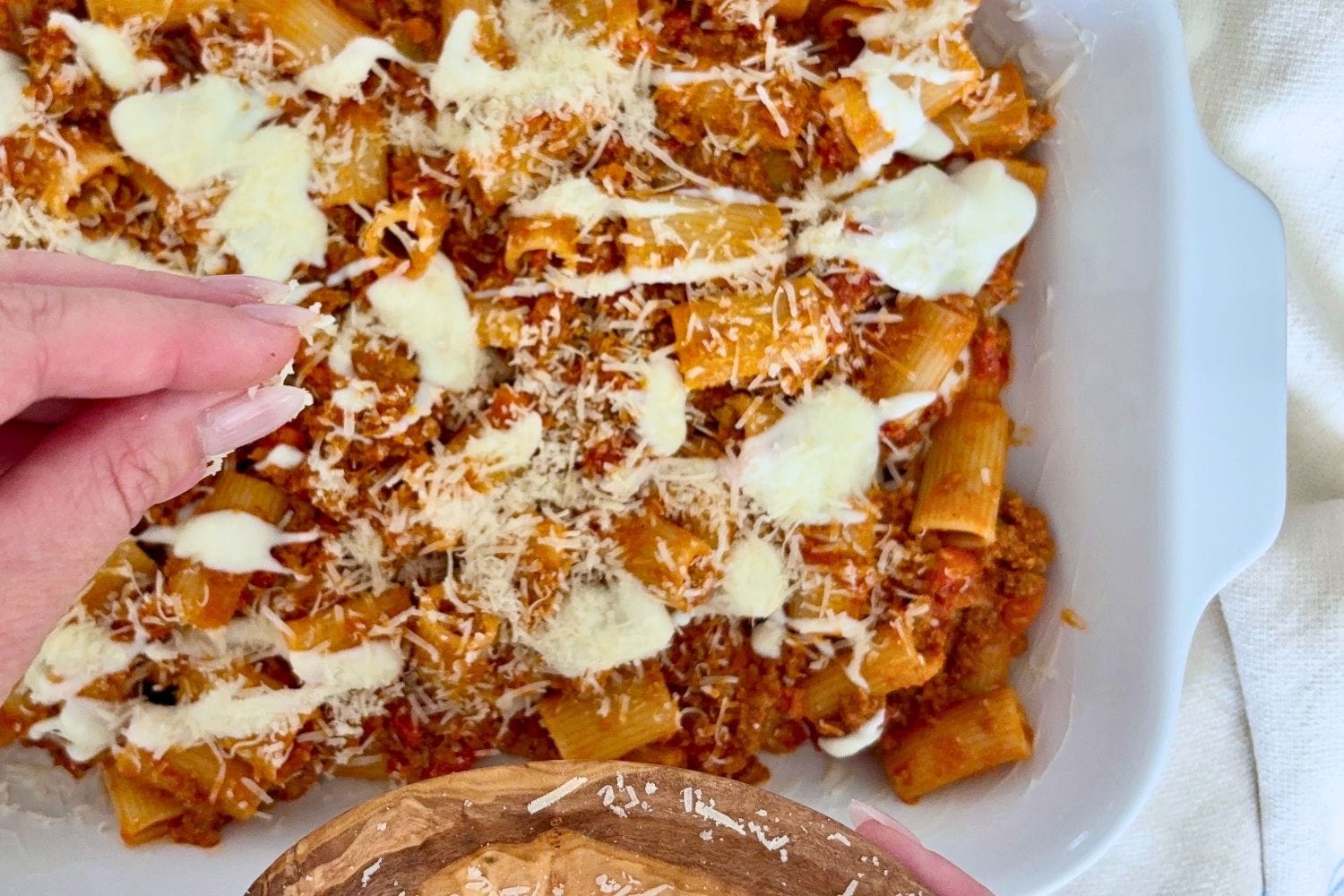 Hand sprinkling grated Parmesan over baked rigatoni with Bolognese and melted cheese in a baking dish