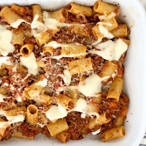Baked rigatoni with Bolognese and béchamel in a white baking dish, topped with melted cheese and grated Parmesan, with a wooden spoon alongside.