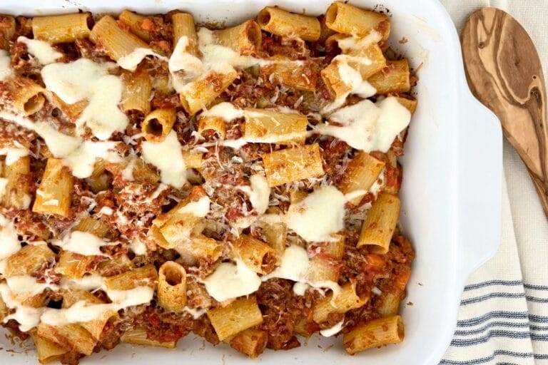Baked rigatoni with Bolognese and béchamel in a white baking dish, topped with melted cheese and grated Parmesan, with a wooden spoon alongside.