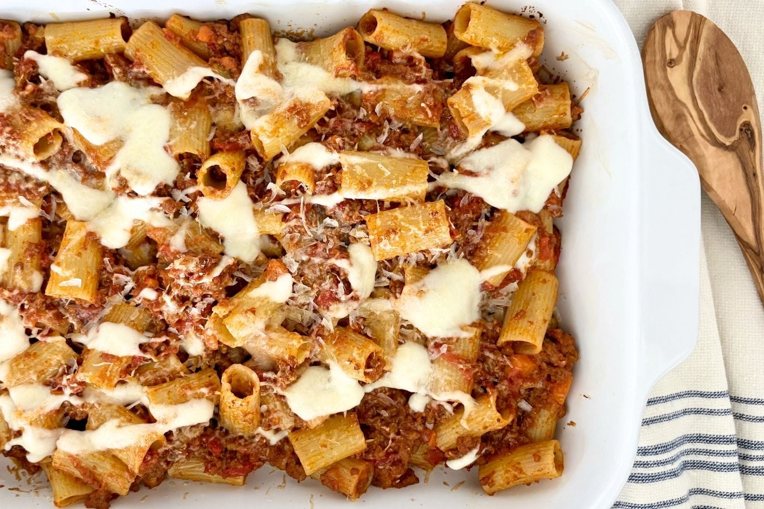 Baked rigatoni with Bolognese and béchamel in a white baking dish, topped with melted cheese and grated Parmesan, with a wooden spoon alongside.