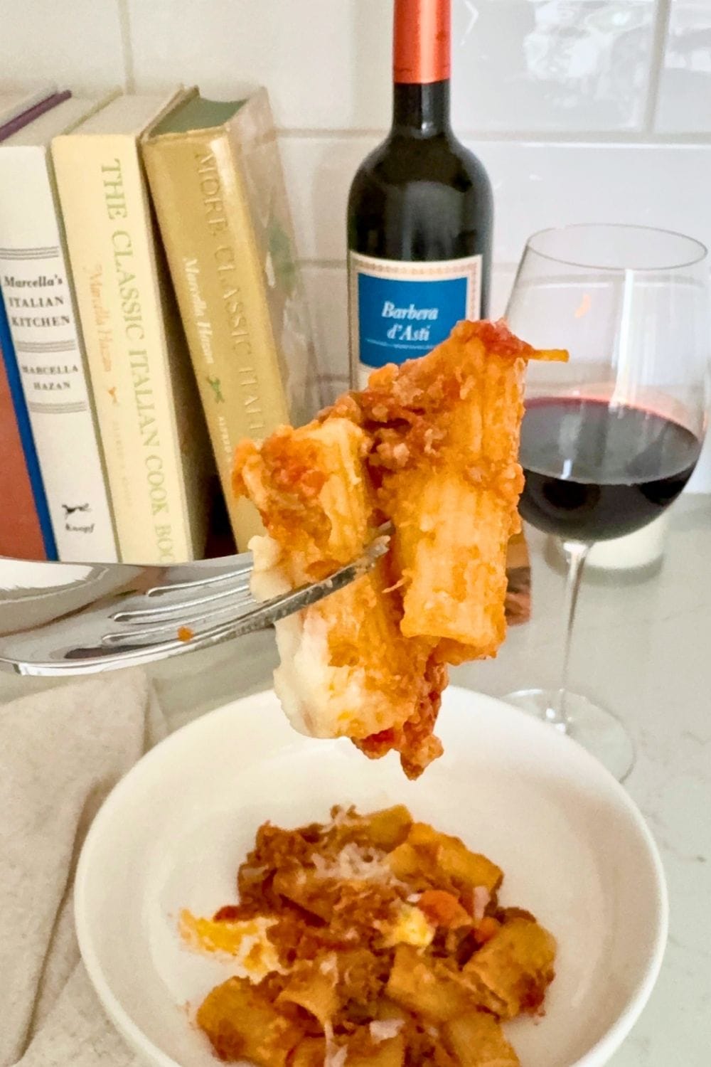 Baked rigatoni with Bolognese on a fork with Barbera wine and Marcella Hazan cookbooks in the background