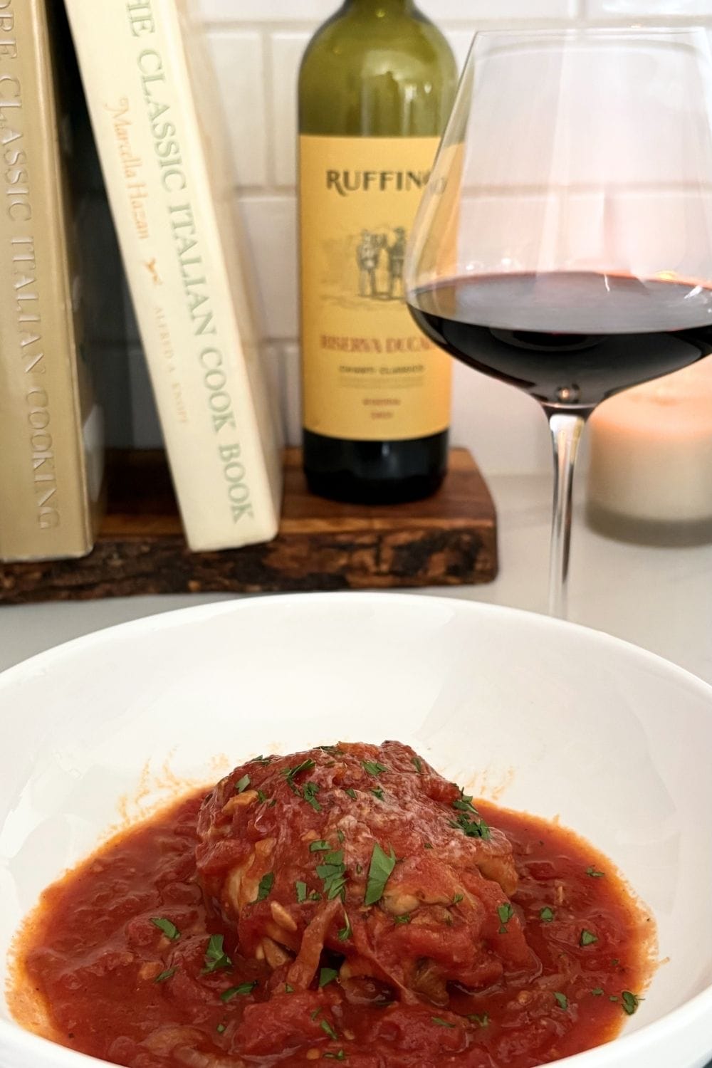 Chicken cacciatore in tomato sauce topped with parsley, served in a white bowl with Chianti and Marcella Hazan cookbook in the background