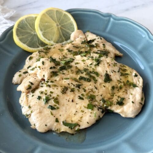 Thin chicken breast fillets with lemon parsley sauce served on a blue scalloped plate with lemon slices on a marble surface.