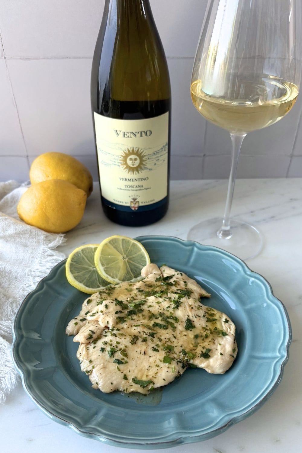 Plated chicken with lemon and parsley served beside a bottle and glass of Vermentino wine with fresh lemons in the background.