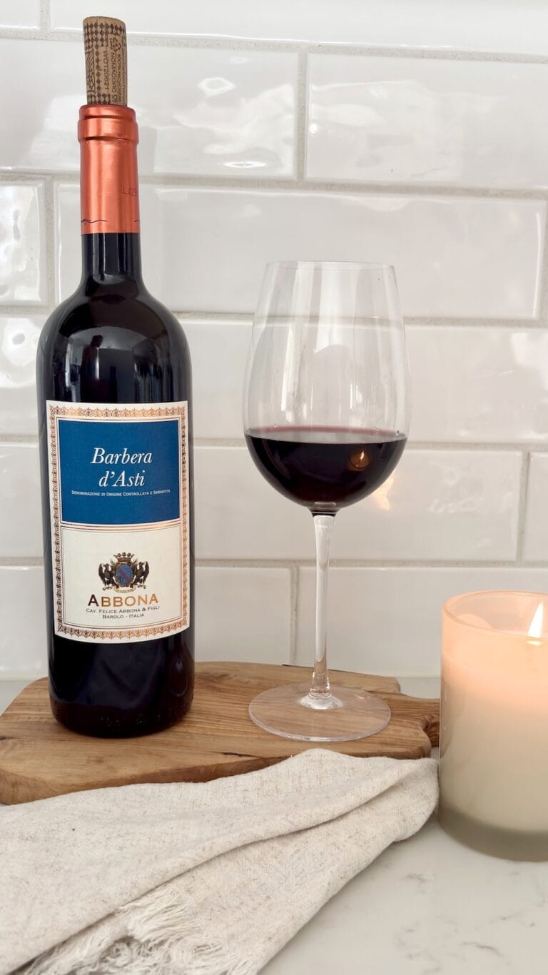 Bottle of Barbera d’Asti red wine beside a glass of red wine on a wooden board with linen cloth and candle against a white tile backsplash.