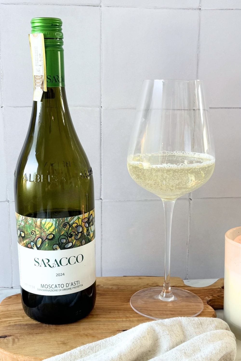 Bottle of Saracco Moscato d’Asti beside a glass of lightly sparkling white wine on a wooden board.