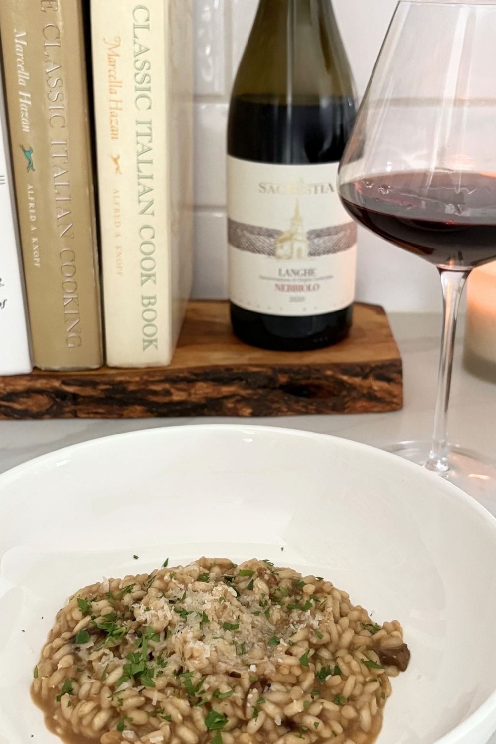 Creamy porcini mushroom risotto in a white bowl with Nebbiolo wine, cookbooks, and candle in the background.