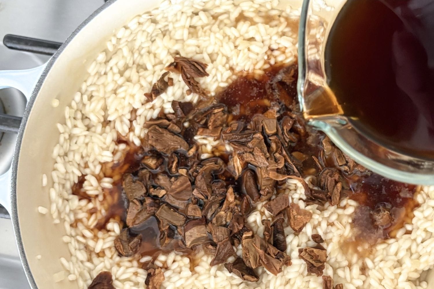 Dark porcini mushroom soaking liquid being poured into a pot of Arborio rice and chopped mushrooms while making risotto.