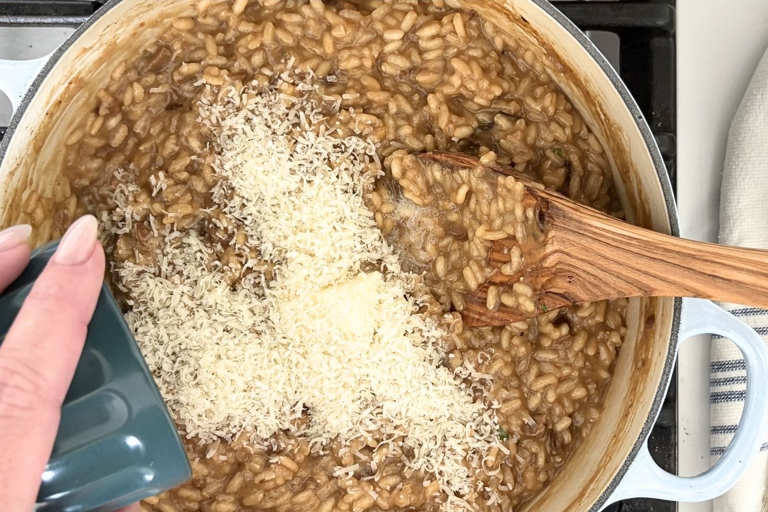Freshly grated Parmesan being added to porcini mushroom risotto in a Dutch oven with wooden spoon.