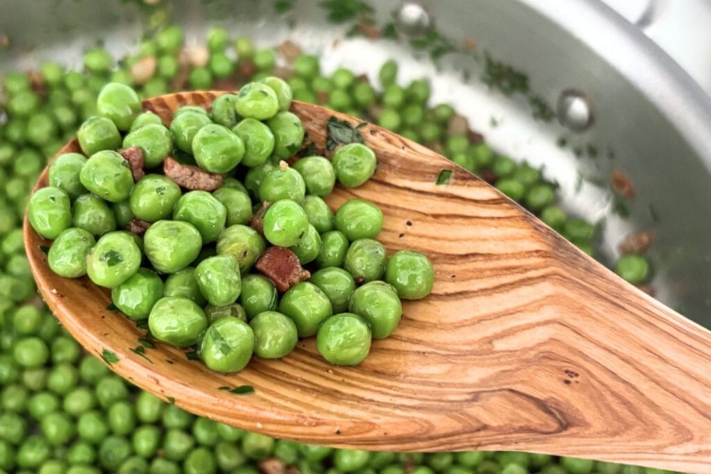 Close-up of sautéed peas with pancetta and parsley on an olive wood spoon over a skillet.