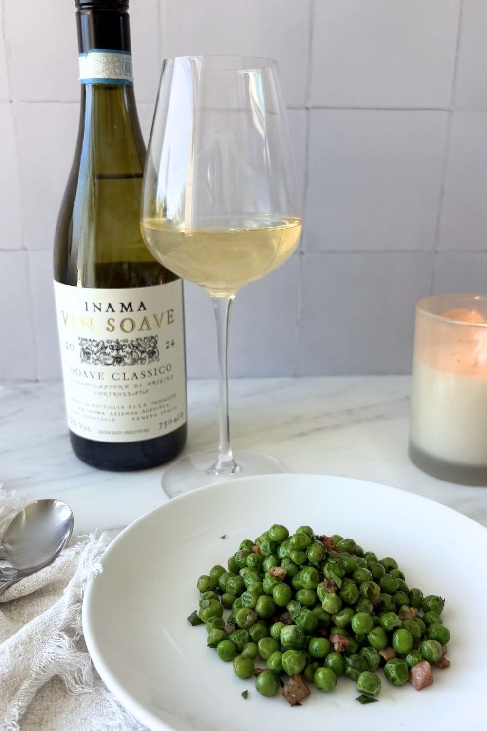 Sautéed peas with pancetta served in a white bowl beside a glass of white wine and bottle of Soave.