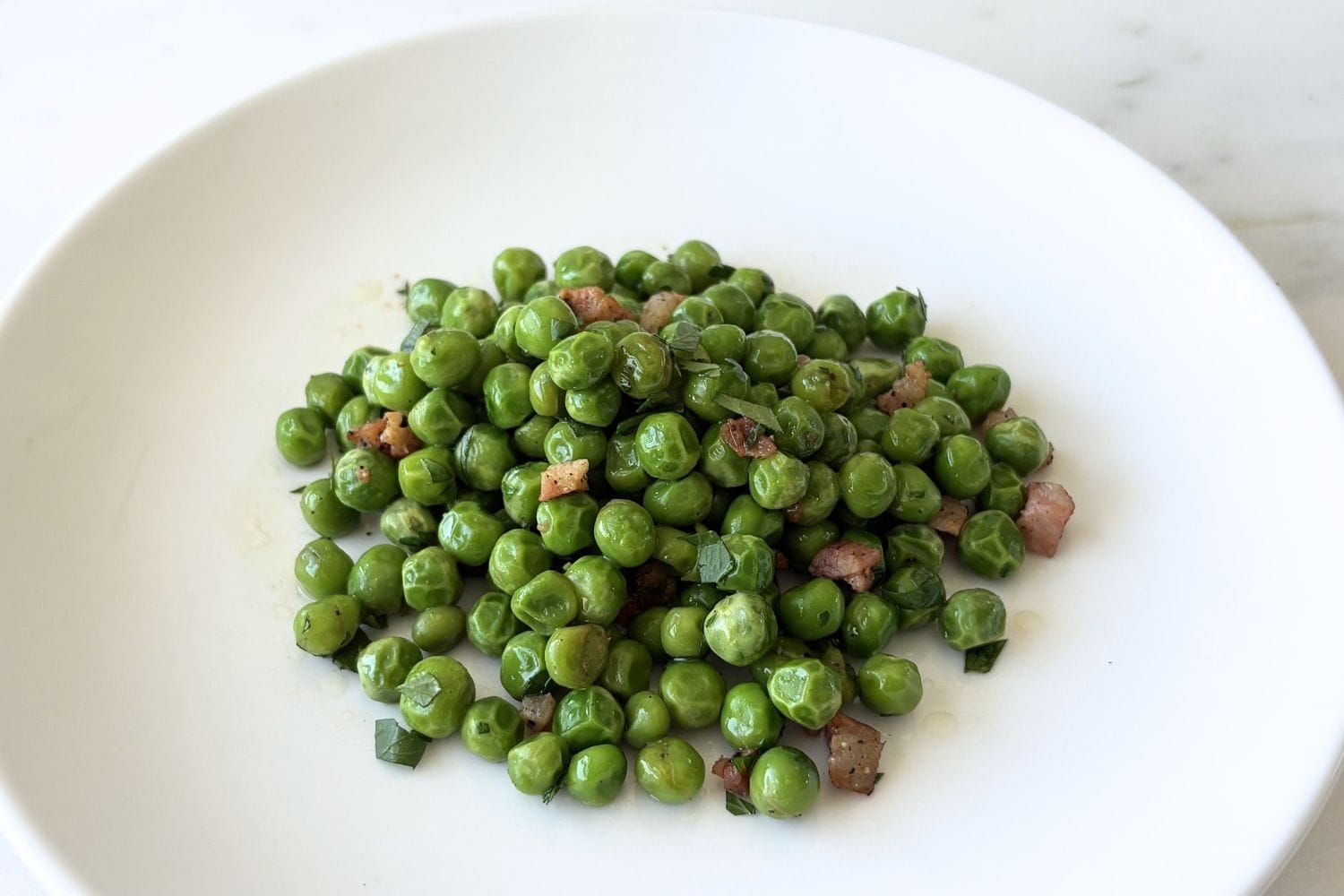 White bowl filled with sautéed peas with pancetta, olive oil, and parsley.