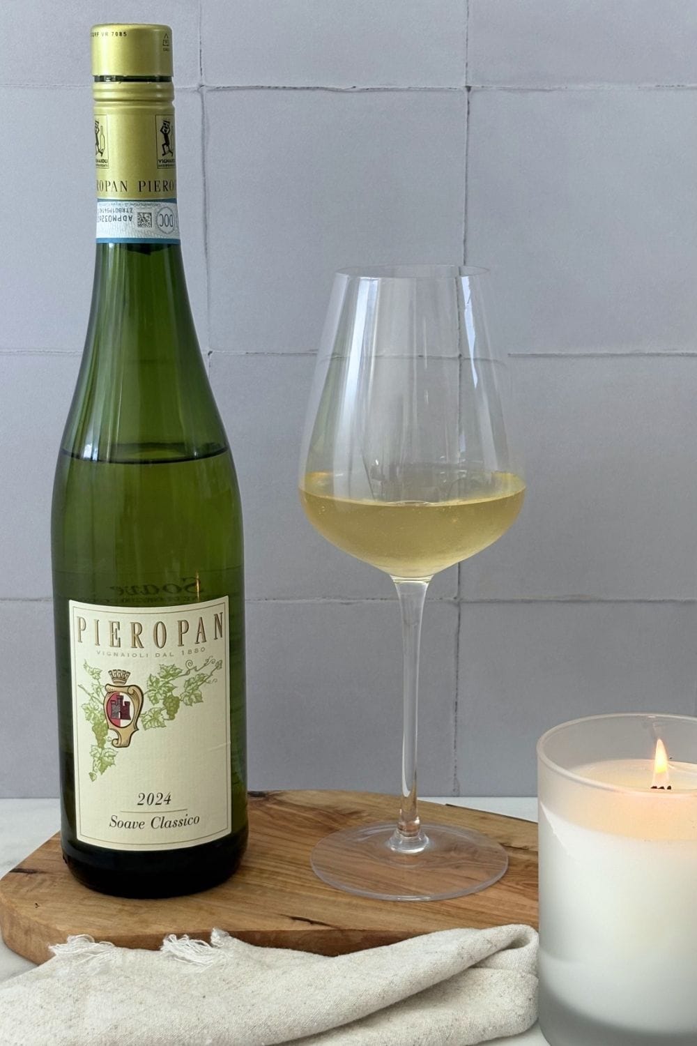 Bottle of 2024 Pieropan Soave Classico beside a glass of pale straw-colored white wine on an olive wood board, styled with a lit white candle, linen napkin, and white tile backdrop.