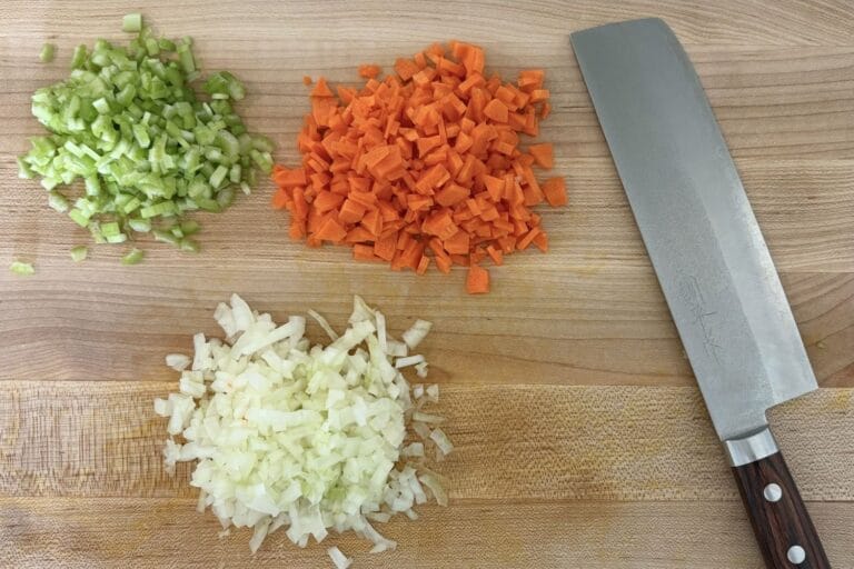 Chopped onion, carrot, and celery arranged on a cutting board beside a chef’s knife for Italian soffritto preparation
