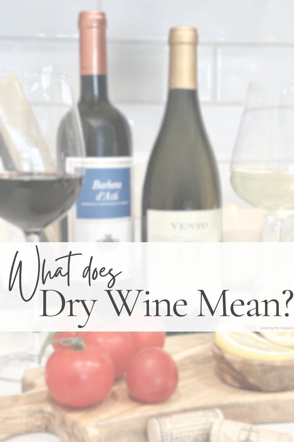 Two wine bottles with red and white wine glasses beside them, styled with tomatoes, lemons, and corks on a wooden board. Text overlay says What Does Dry Wine Mean?