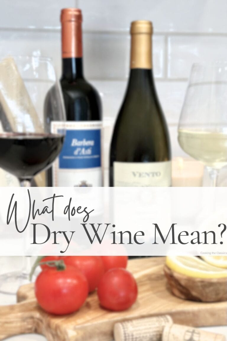 Two wine bottles with red and white wine glasses beside them, styled with tomatoes, lemons, and corks on a wooden board. Text overlay says What Does Dry Wine Mean?