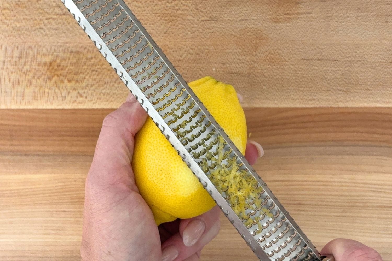 Hand zesting a fresh lemon with a microplane over a wooden cutting board.