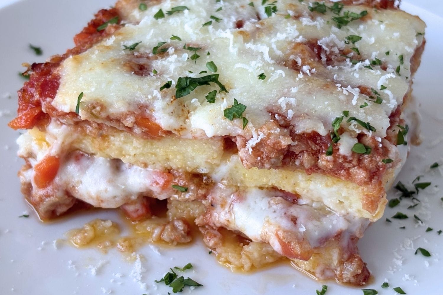 Close-up of baked polenta layered with Bolognese sauce, béchamel, and parmesan cheese, topped with parsley and served warm on a white plate.