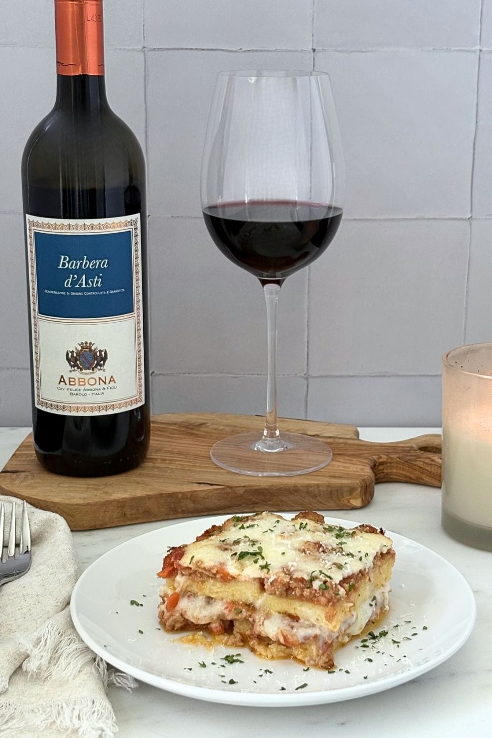 Slice of baked polenta with Bolognese and béchamel paired with a glass and bottle of Barbera wine on an olive wood board beside a candle.