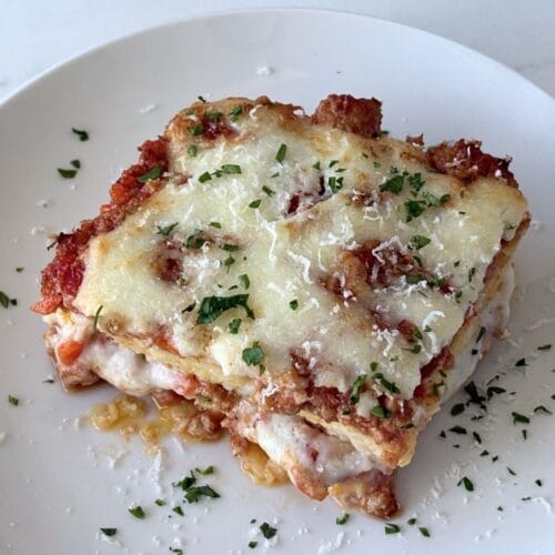Baked polenta with layers of Bolognese, béchamel, and parmesan served on a white plate with parsley scattered over the top.