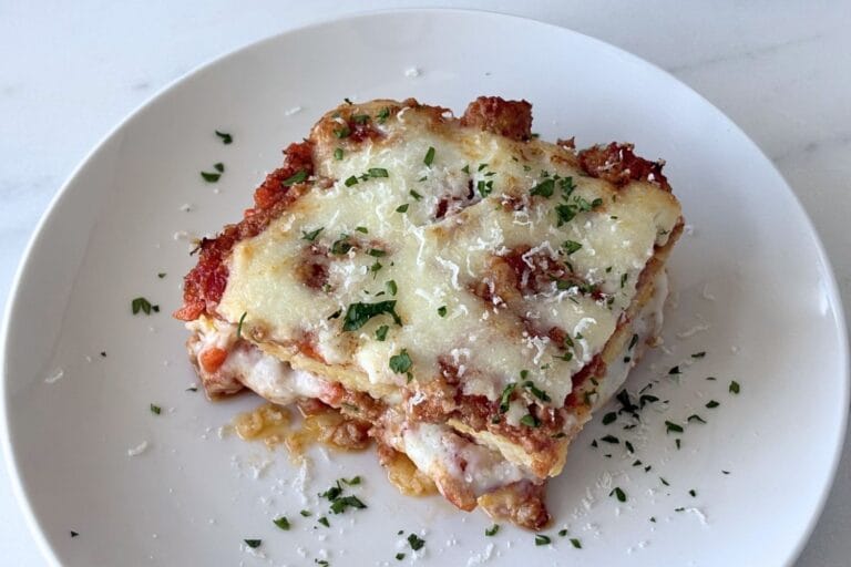 Baked polenta with layers of Bolognese, béchamel, and parmesan served on a white plate with parsley scattered over the top.