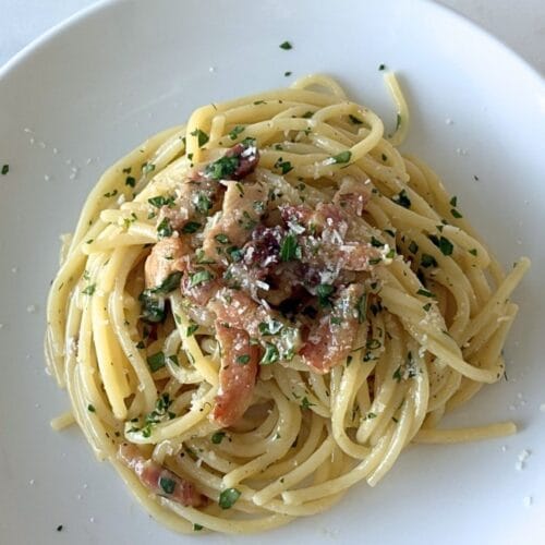 White bowl of Marcella Hazan’s carbonara sauce served with spaghetti, pancetta, parsley, black pepper, and grated cheese on a marble surface.