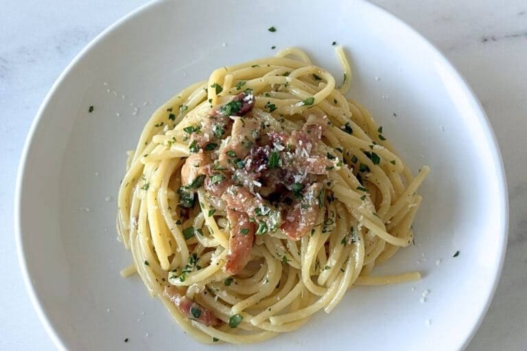 White bowl of Marcella Hazan’s carbonara sauce served with spaghetti, pancetta, parsley, black pepper, and grated cheese on a marble surface.