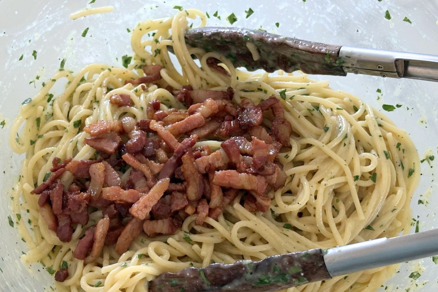 Spaghetti tossed with egg and cheese sauce in a large bowl with crisp pancetta added on top before mixing.