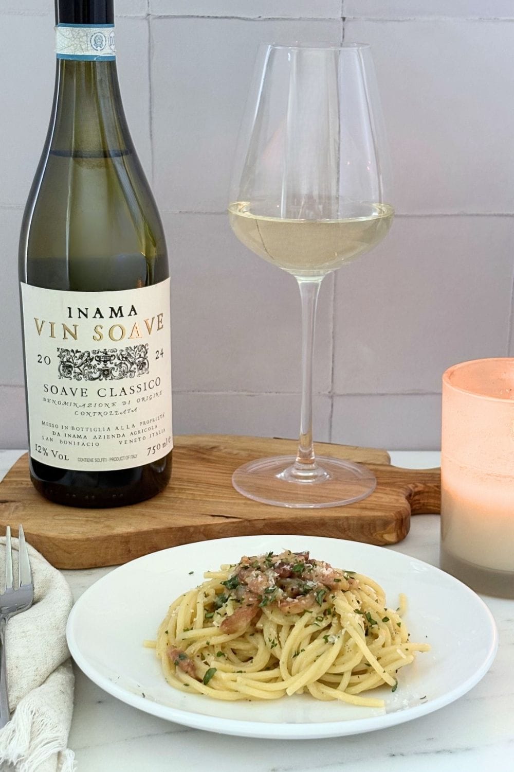 Plate of Marcella Hazan’s carbonara sauce served with a glass and bottle of Soave Classico on a marble countertop beside a candle.