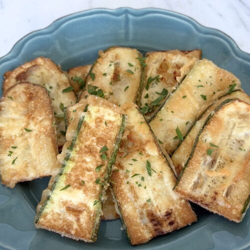 Golden fried zucchini slices with a thin flour batter served on a blue plate with chopped parsley.
