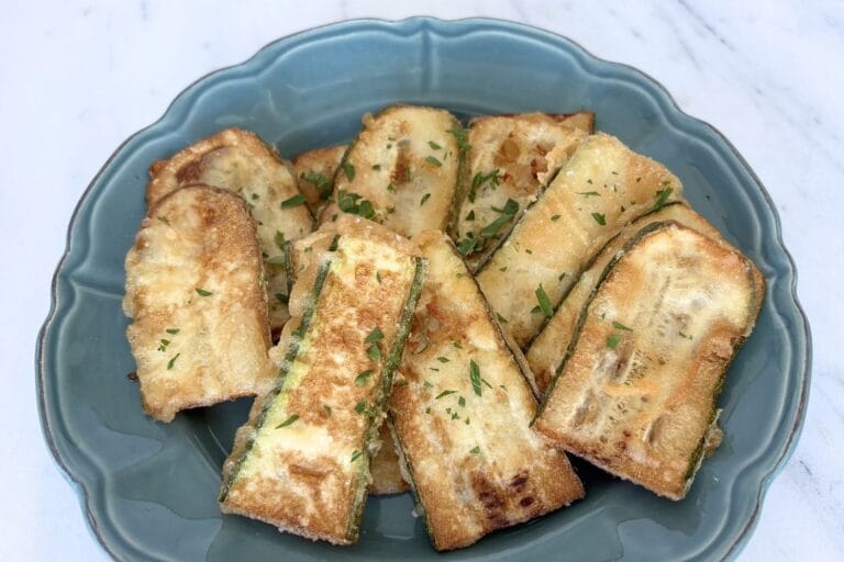 Golden fried zucchini slices with a thin flour batter served on a blue plate with chopped parsley.