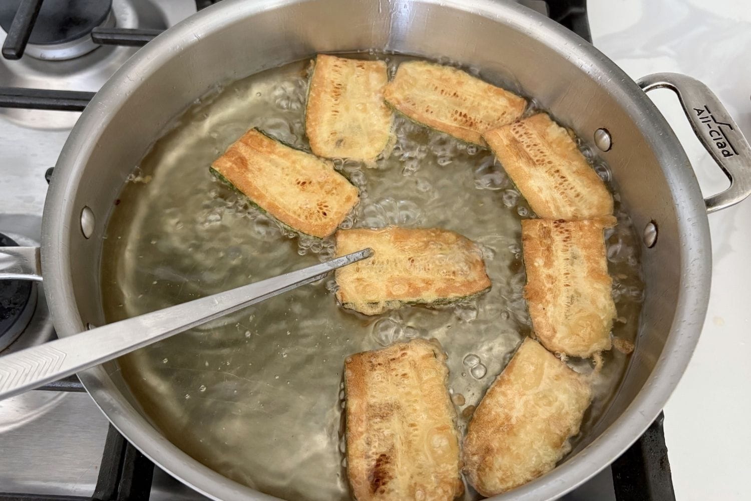 Thin battered zucchini slices frying in deep vegetable oil in a stainless steel pan until golden brown.