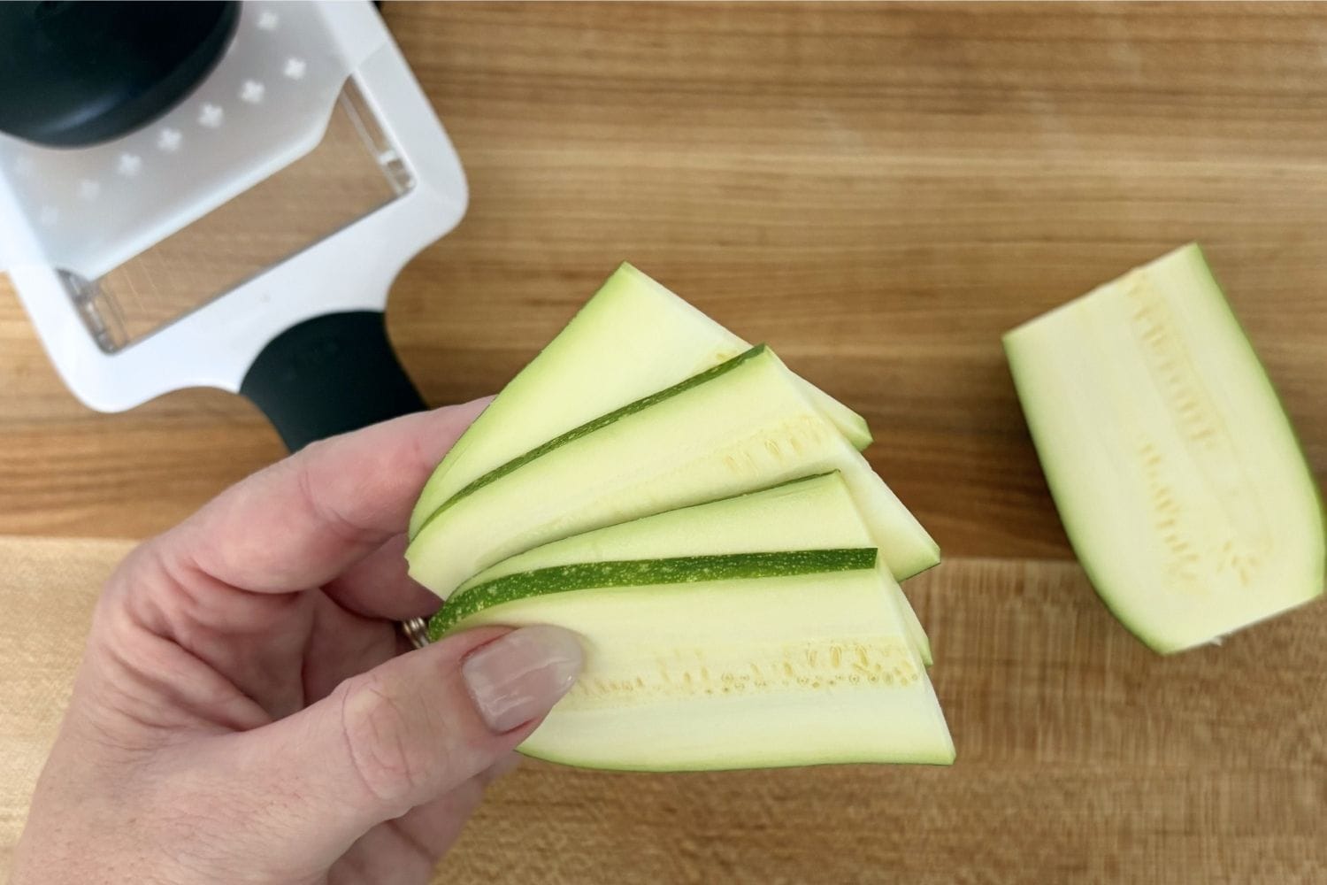 Thin zucchini slices cut lengthwise beside a handheld mandoline slicer on a wooden cutting board for Marcella Hazan’s fried zucchini recipe.