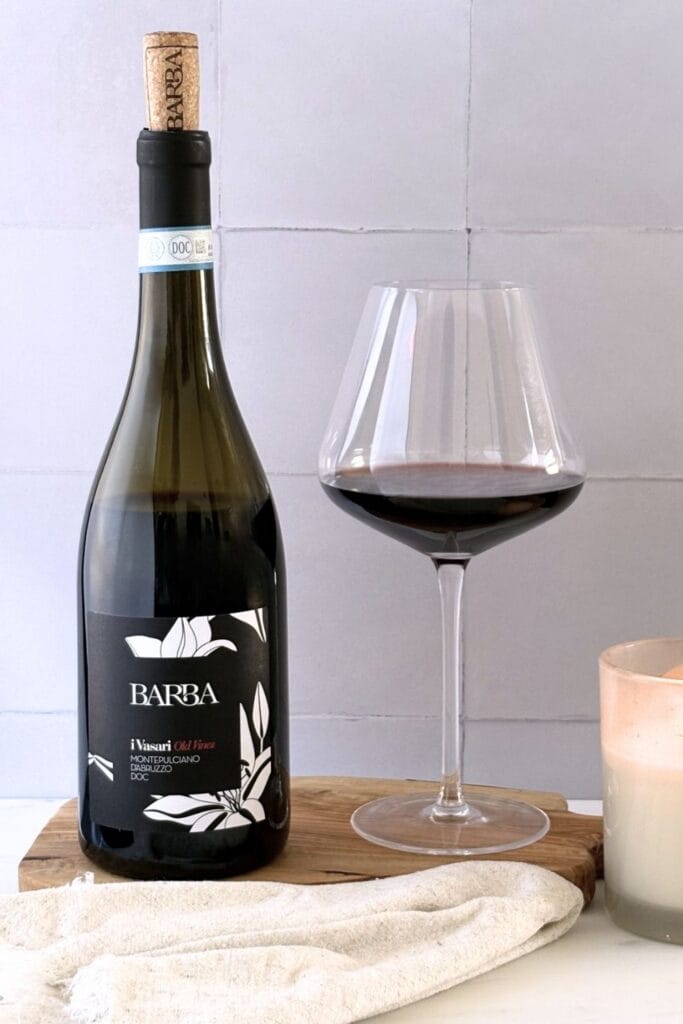 Bottle of Montepulciano d’Abruzzo red wine with a glass of red wine on an olive wood board beside a candle and linen cloth.