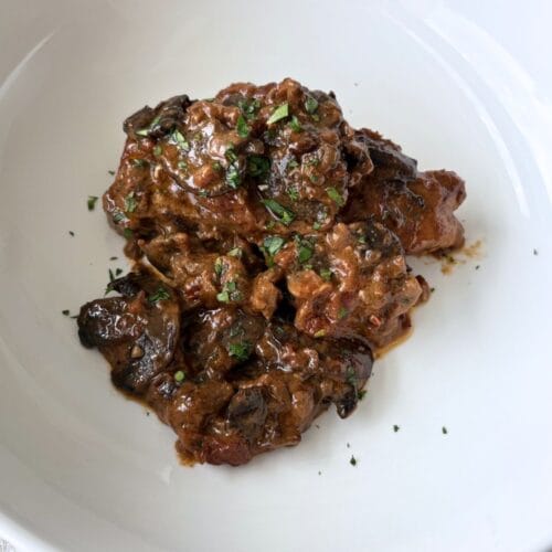 Braised pork chops with tomatoes, cream, and porcini mushrooms served in a white bowl, finished with chopped parsley