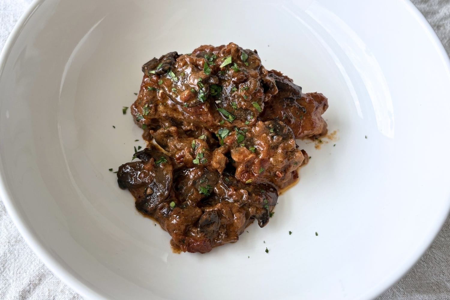Braised pork chops with tomatoes, cream, and porcini mushrooms served in a white bowl, finished with chopped parsley