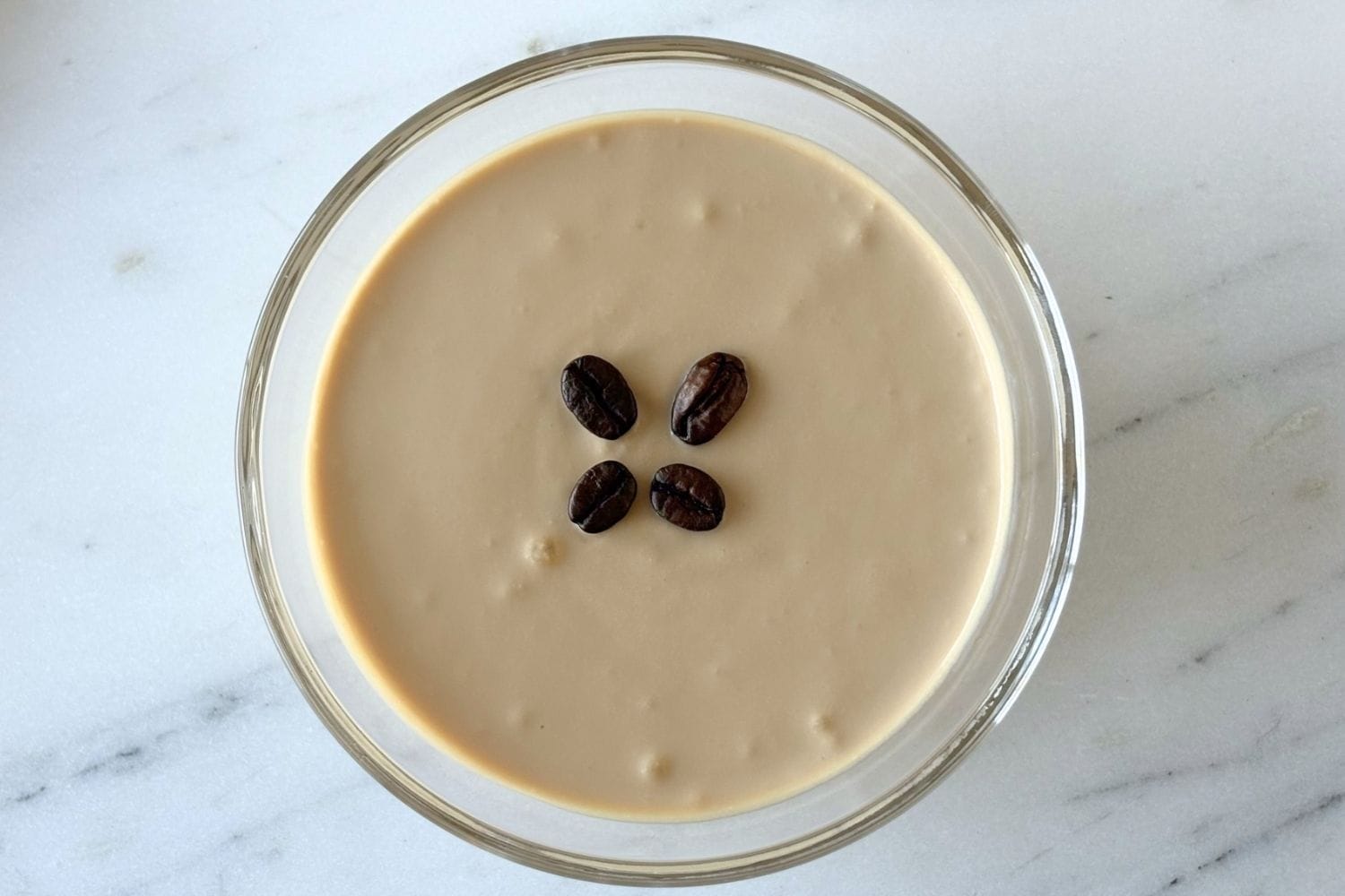 Top-down view of Marcella Hazan’s ricotta and coffee cream dessert in a glass bowl topped with four coffee beans on a marble surface.