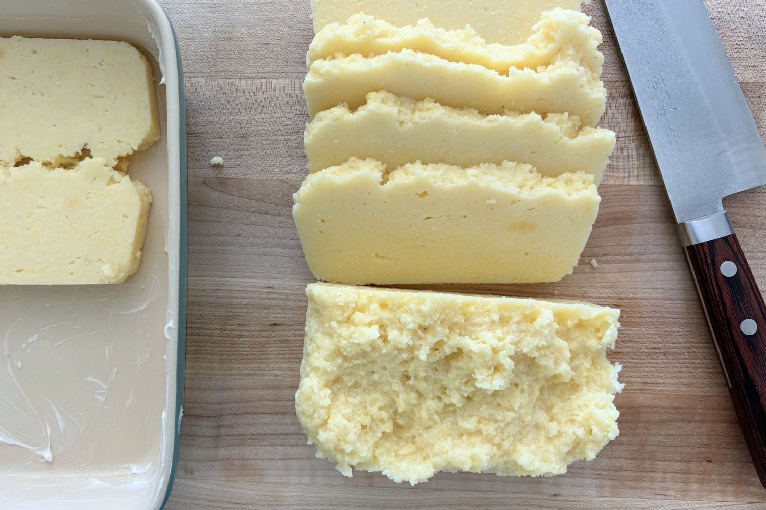Sliced chilled polenta on a wooden cutting board beside a chef’s knife, showing the texture of leftover polenta before layering into baked polenta with Bolognese.