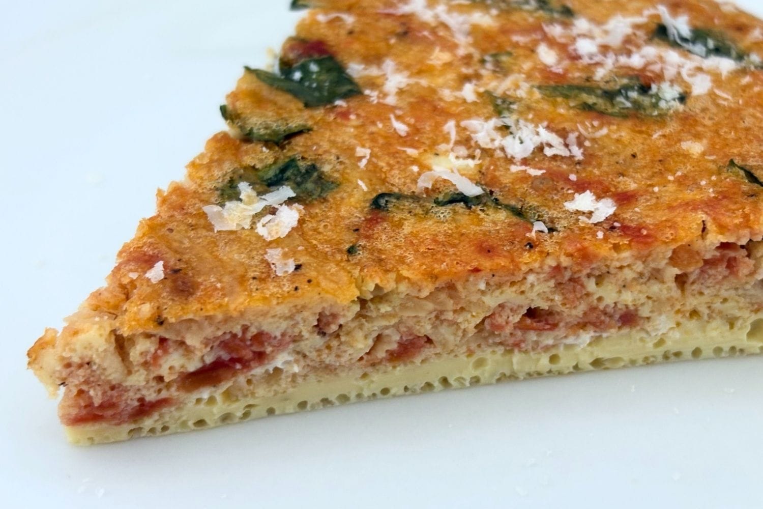 Close-up of a slice of Marcella Hazan's tomato, onion, and basil frittata topped with freshly grated Parmigiano-Reggiano. The baked egg is golden brown with pieces of tomato and basil visible throughout.