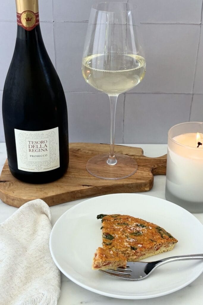 A plated slice of tomato, onion, and basil frittata served with a bottle and glass of Prosecco on an olive wood board. A white candle and linen napkin complete the table setting.