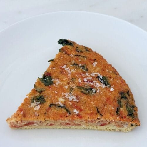 A slice of tomato, onion, and basil frittata on a white plate, finished with freshly grated Parmigiano-Reggiano. The golden baked egg is filled with tomatoes and basil.