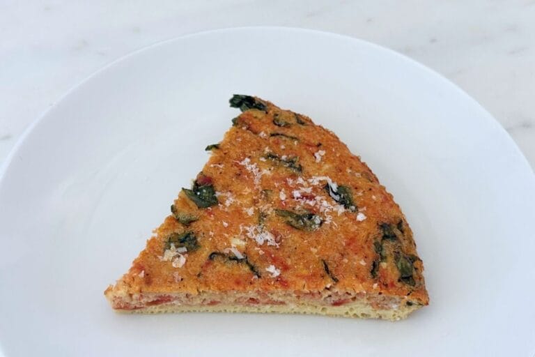 A slice of tomato, onion, and basil frittata on a white plate, finished with freshly grated Parmigiano-Reggiano. The golden baked egg is filled with tomatoes and basil.