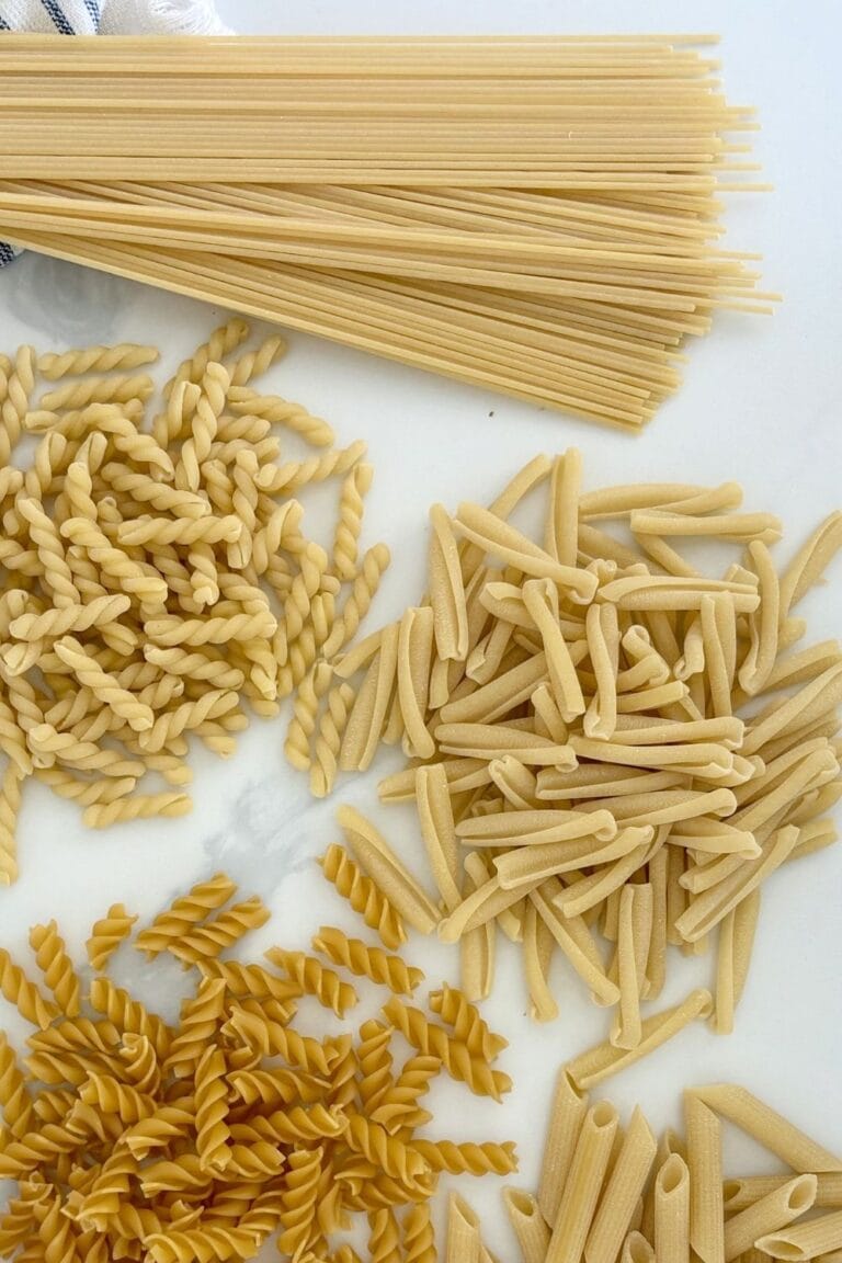 Assorted dried pasta shapes including spaghetti, fusilli, casarecce, and penne arranged on a white marble surface.