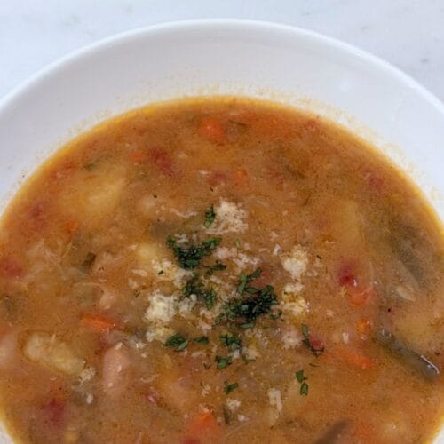 Minestrone alla Romagnola in a white bowl topped with grated Parmigiano-Reggiano and chopped parsley on a marble surface.