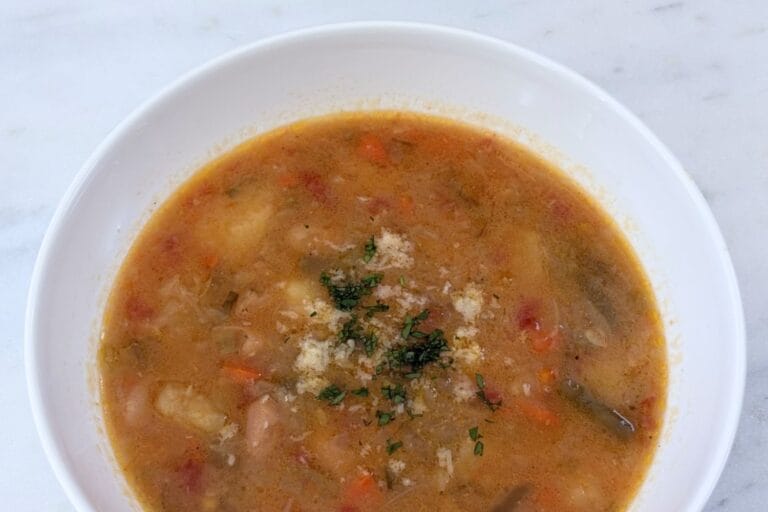 Minestrone alla Romagnola in a white bowl topped with grated Parmigiano-Reggiano and chopped parsley on a marble surface.