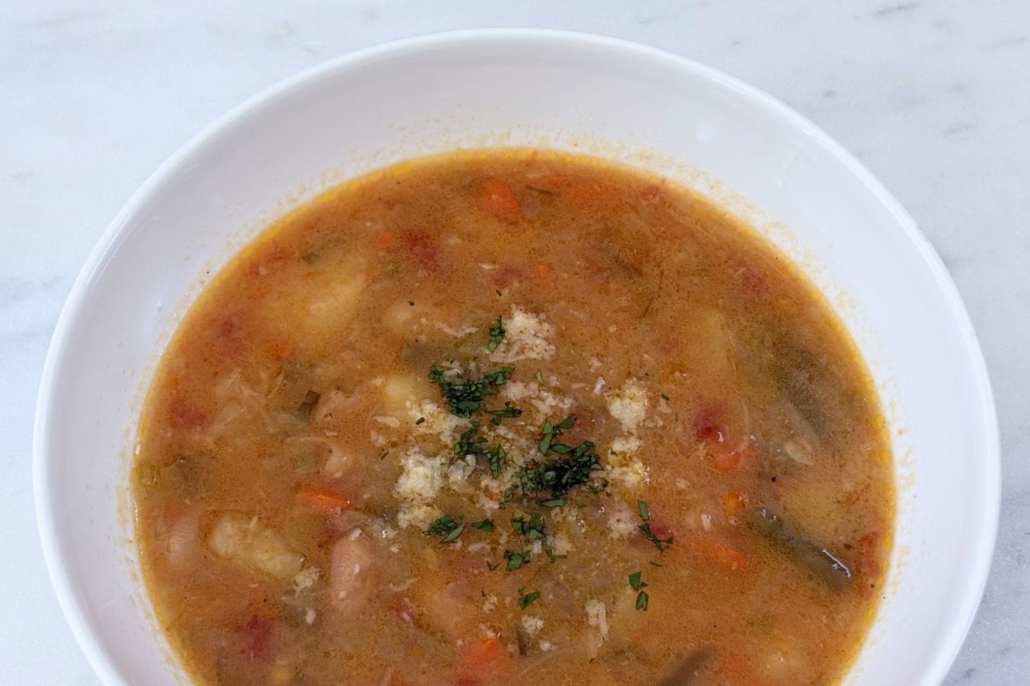 Minestrone alla Romagnola in a white bowl topped with grated Parmigiano-Reggiano and chopped parsley on a marble surface.