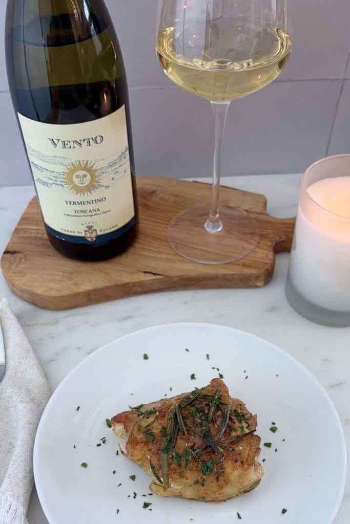 Pan-roasted chicken with rosemary and garlic served alongside a glass and bottle of Vermentino on a wooden board.