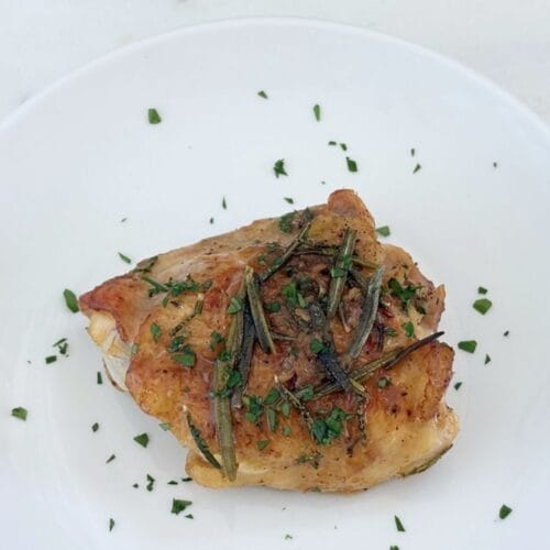 A single bone-in, skin-on chicken thigh served on a white plate with rosemary, pan juices, and chopped parsley.
